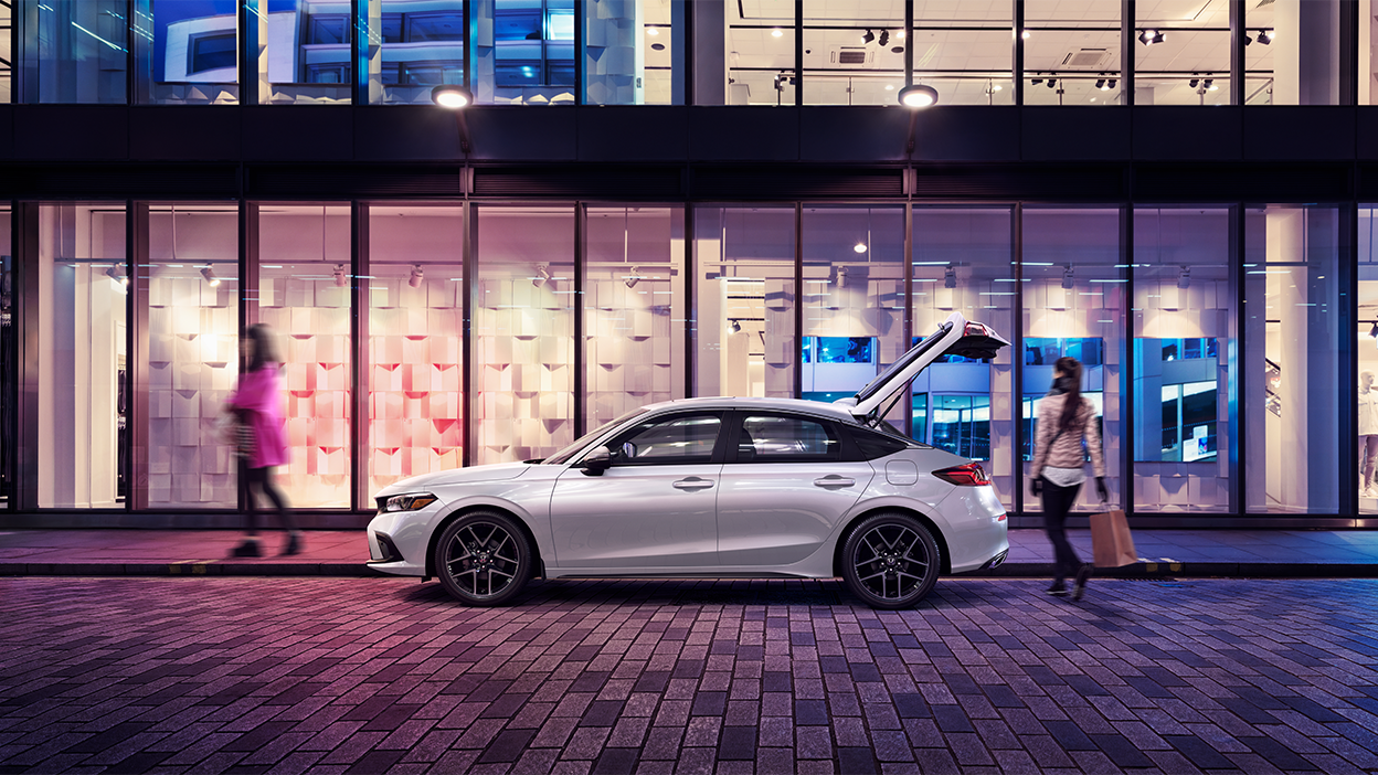 Side view of white Hatchback parked outside a retail store with the hatch door open. A couple shoppers walk past it.