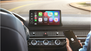 Medium closeup of touchscreen on home page, seen over the shoulder of driver and front passenger. Front passenger holds phone with app open. 