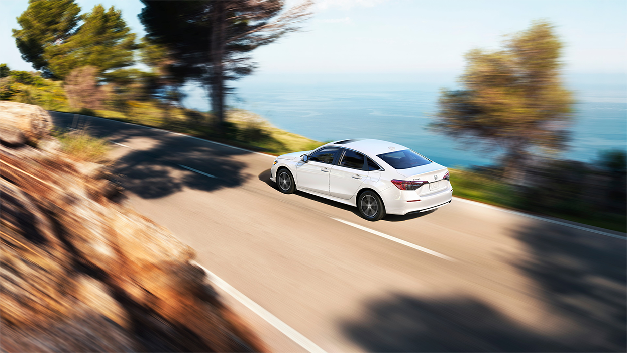 3/4 rear bird’s eye view of white Civic Sedan driving on an oceanside highway in the day.