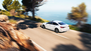 3/4 rear bird’s eye view of white Civic Sedan driving on an oceanside highway in the day. 