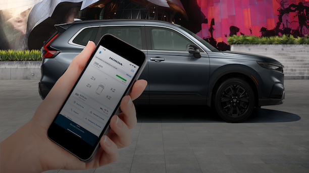 Sideview of grey CR-V parked in front of a graffitied wall. Hand in foreground holds a smartphone with the HondaLink™ app. 