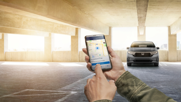 Hands using HondaLink app on phone. Grey odyssey parked in the background.