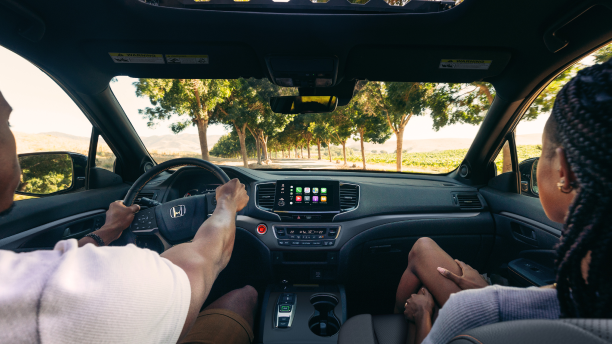 Interior view of the front dashboard seen over the shoulder of driver and passenger. Through the windshield we see they’re drying on a treelined road. 