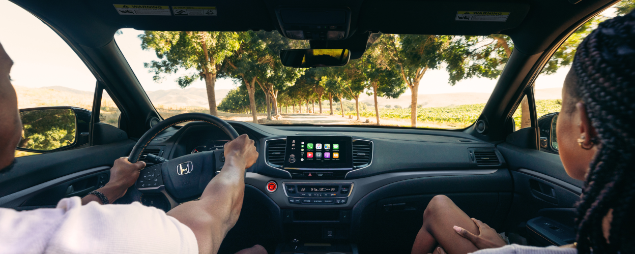 Interior view of the front dashboard seen over the shoulder of driver and passenger. Through the windshield we see they’re drying on a treelined road.