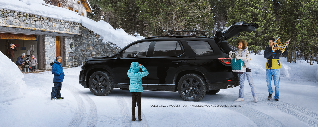 Plan latéral de trois quarts d’un Pilot TrailSport noir stationné dans l’entrée enneigée d’un chalet moderne avec la porte de garage ouverte. Trois générations d’une même famille l’entourent. Maman et papa déchargent le hayon ouvert.