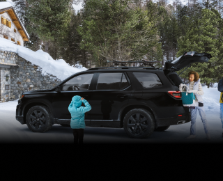 Plan latéral de trois quarts d’un Pilot TrailSport noir stationné dans l’entrée enneigée d’un chalet moderne avec la porte de garage ouverte. Trois générations d’une même famille l’entourent. Maman et papa déchargent le hayon ouvert.