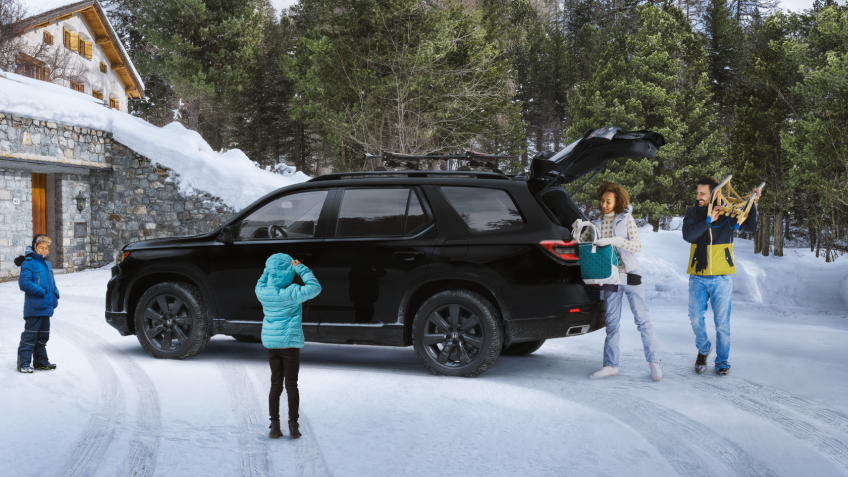 Plan latéral de trois quarts d’un Pilot TrailSport noir stationné dans l’entrée enneigée d’un chalet moderne avec la porte de garage ouverte. Trois générations d’une même famille l’entourent. Maman et papa déchargent le hayon ouvert.