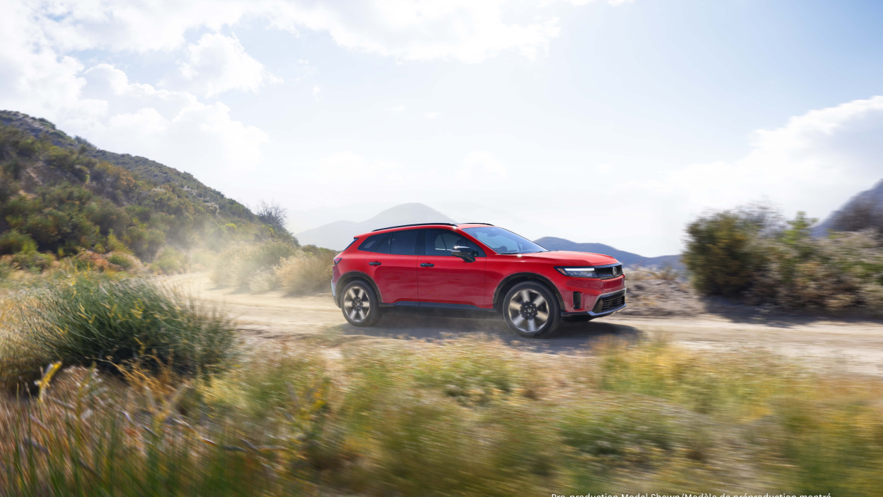 Wide 3/4 side view of red Prologue driving on desert road, kicking up dust. 