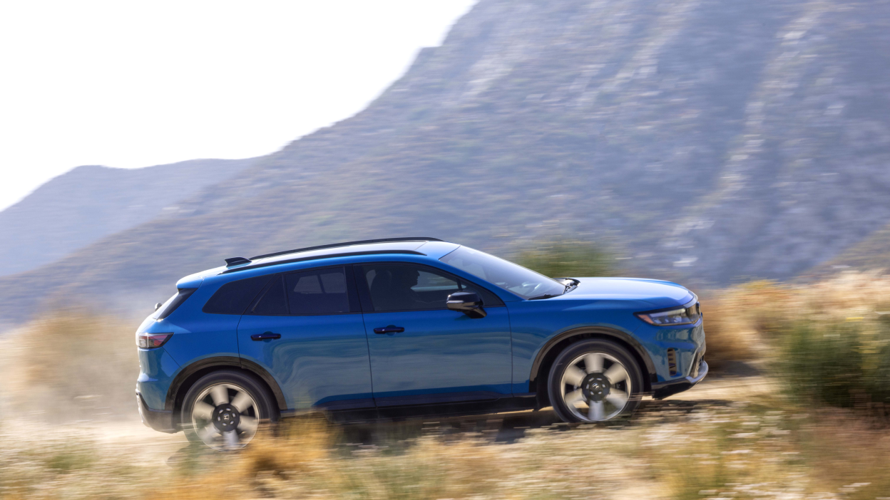 Wide side view of blue Prologue driving on desert road, kicking up dust. 