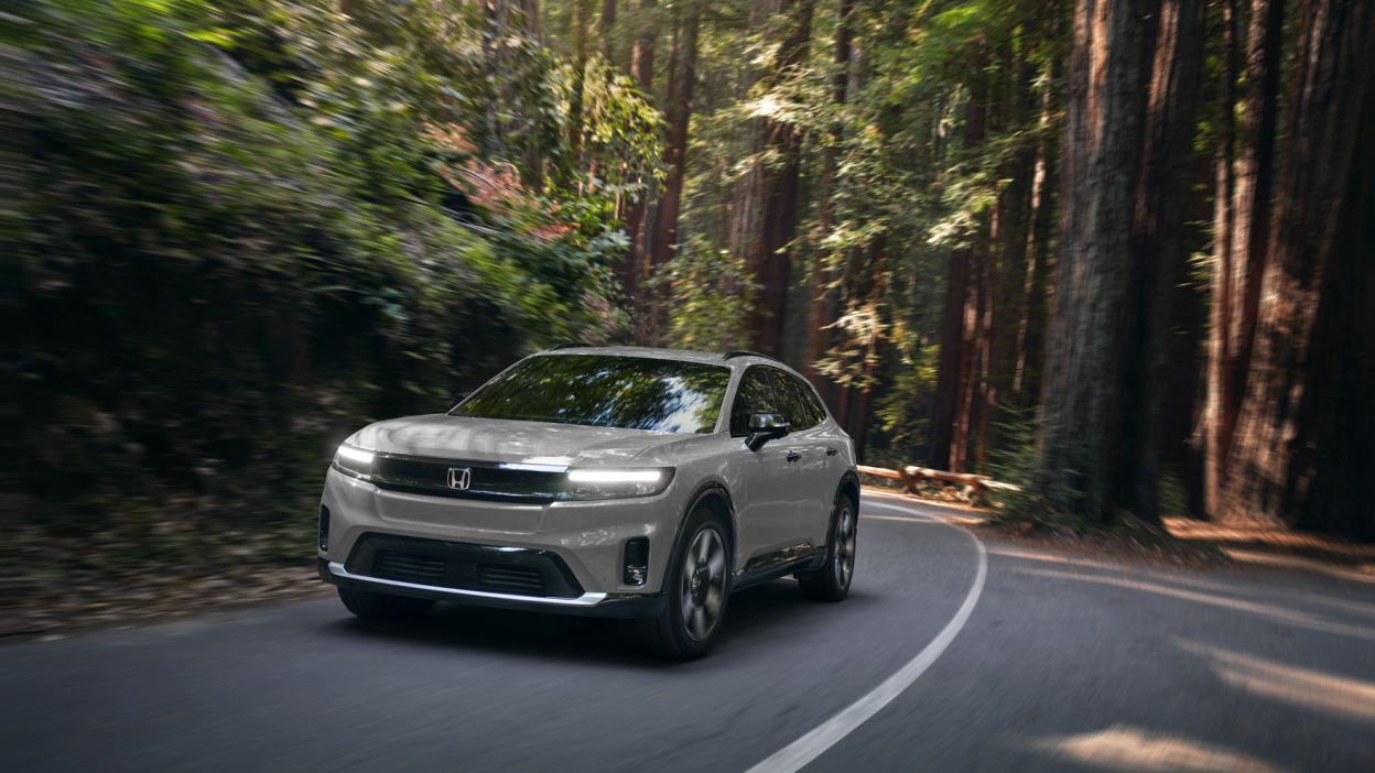 3/4 front view of white Prologue taking a turn on a paved road in a temperate forest.
