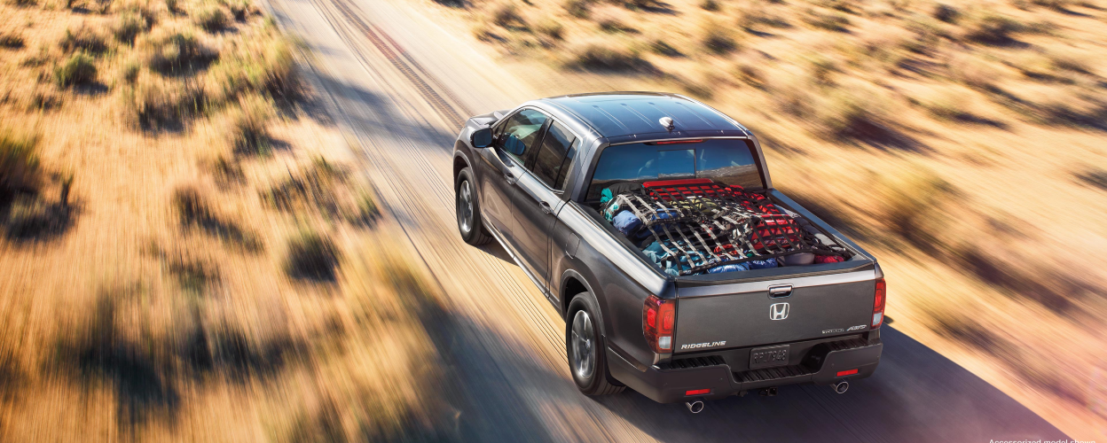 3/4 rear bird’s eye view of grey Ridgeline driving on desert road. Truck-bed loaded with stuff.