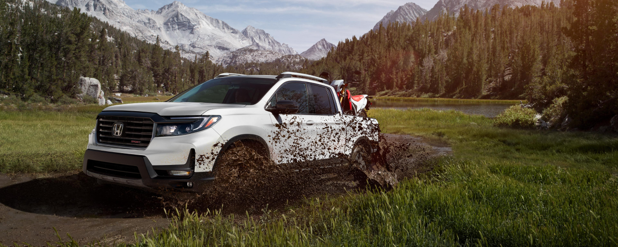 3/4 front side view of white Ridgeline with motocross bikes in truck-bed, barrelling thru mud puddle.